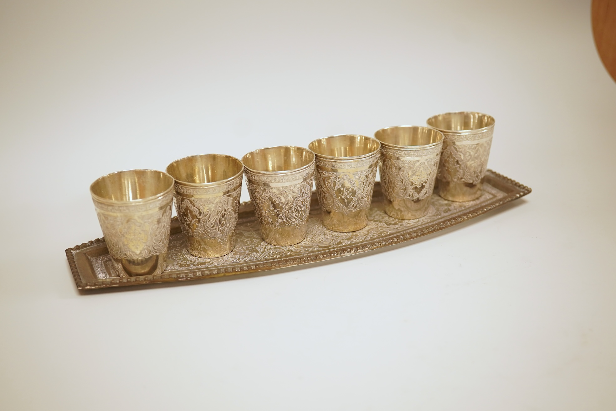 A set of six 20th century Middle Eastern white metal tots, 42mm and matching tray, 25.2cm, all engraved with geometric foliate decoration.